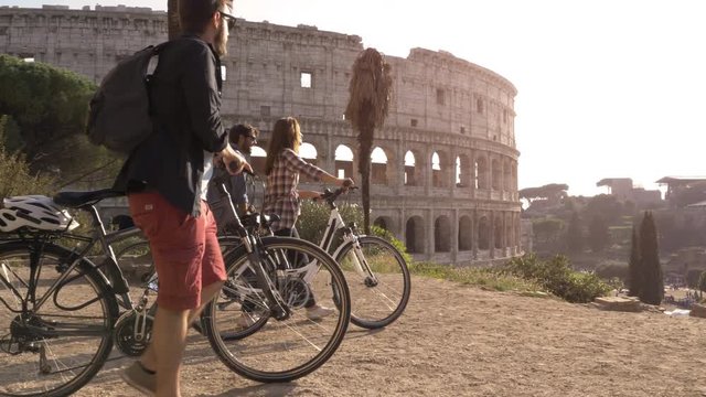 Three happy young friends tourists with bikes and backpacks at Colosseum in Rome arriving on hill at sunset with trees slow motion steadycam