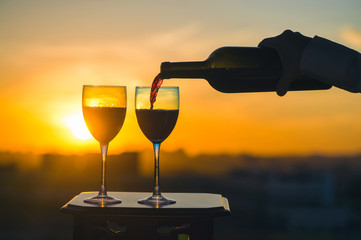 Female hand with bottle pours red wine into glasses on blurred night city background. Service on the roof of the restaurant