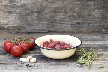 Cut into pieces of beef meat in a light bowl, tomatoes on a branch, garlic, thyme, rosemary, on a wooden background in rustic style