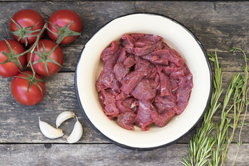 Cut into pieces of beef meat in a light bowl, tomatoes on a branch, garlic, thyme, rosemary, on a wooden background in rustic style