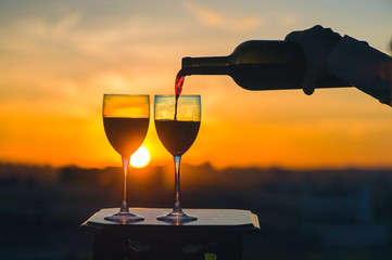 Female hand with bottle pours red wine into glasses on blurred night city background. Service on the roof of the restaurant