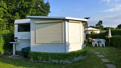 Caravan mit einer festen Veranda aus Markisenstoff, Glasschiebefenster und Rollläden auf einem deutschen Campingplatz. Komfortabel und gut gepflegt. Die Jalousien sind geschlossen.
