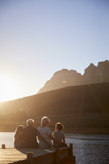 Grandchildren With Grandparents Sitting On Wooden Jetty By Lake