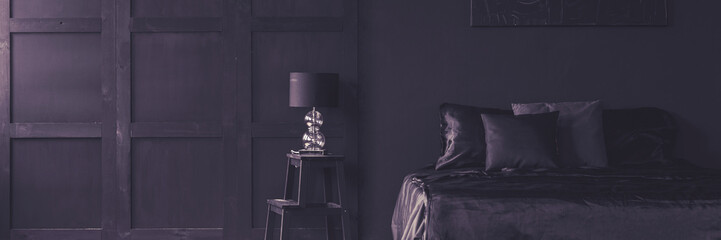Desk lamp on a ladder side able and a cozy bed with satin sheets and pillows in an elegant, dark purple apartment interior