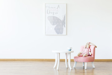 Plush toy on pink chair at table in white girl's room interior with butterfly poster on the wall. Real photo