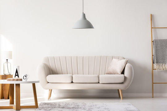 Real photo of a beige sofa standing in a simple living room interior under a lamp and next to a table and ladder