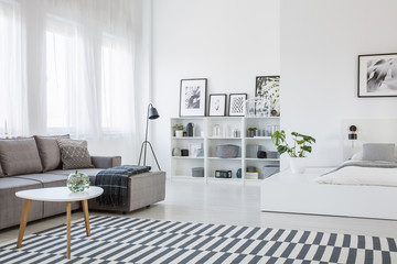 Table on patterned carpet in open space interior with white bed and grey corner sofa. Real photo