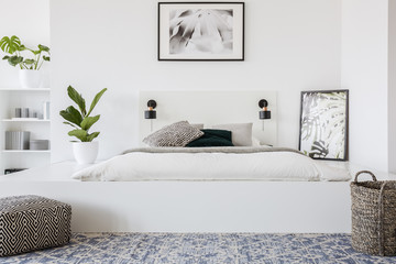 Basket and pouf in front of bed on platform in bright bedroom interior with plant and poster. Real photo