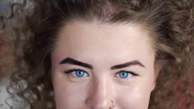 Video portrait of curly woman that opes her blue eyes and smiles, attractive woman with piercing in her nose, girl with big blue eyes