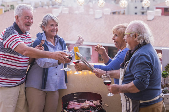 Four Active Senior Retired Have Fun In The Terrace At Home Cooking Some Bbq Everybody Smiles And Stay Together In Friendship Under A Nice Sunny Day.  Vacation And Relationship People Concept