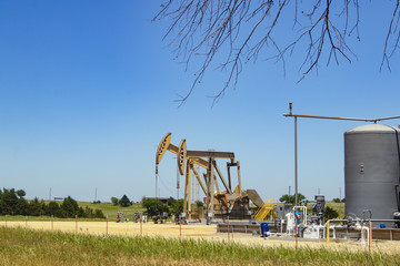 Two pump jacks at a pumping station with tanks and machinery with some farm buildings on the...
