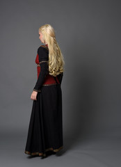 Fototapeta premium full length portrait of pretty blonde lady wearing a red and black fantasy medieval gown. standing pose on grey background.