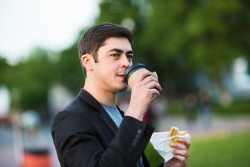 Closeup portrait of businessman holding pastry and drinking coffee at green park background.