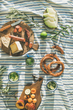 Flat-lay of summer picnic set with fruit, cheese, sausage, bagels and lemonade over striped blanket, top view, vertical