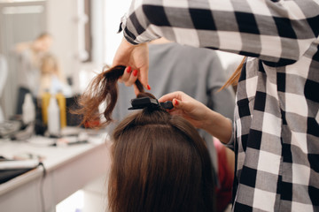 Naklejka premium Hairdresser woman straightens her hair and makes styling, close-up of curling iron in salon
