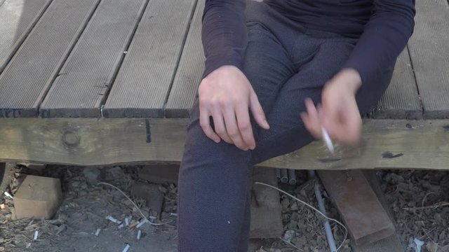 A man holds a cigarette in his hands and nervously smokes, sitting on wooden boards