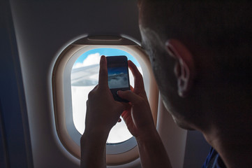 Young passenger man taking photo of the aircraft wing and clouds while sitting inside the airplane during flight.