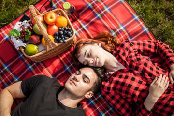 Loving young couple lying on picnic blanket near basket full of fruits enjoying sunny day