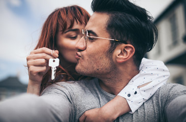 Loving couple with keys to their new home hugging and looking at camera taking selfie