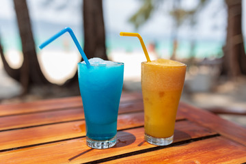 Two glasses with a blue and orange cocktail stand on a wooden table under the rays of the sun.