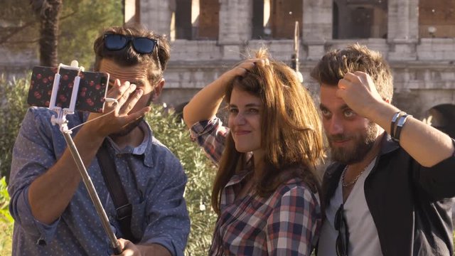 Three happy young friends tourists at Colosseum in Rome taking funny selfies with smartphone stick making faces on hill at sunset with trees slow motion steadycam