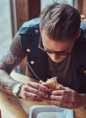 Portrait of a hungry hipster guy with a stylish haircut and beard sits at a table, decided to dine at a roadside cafe, eating a hamburger.