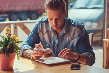 Portrait of a hungry hipster guy with a stylish haircut and beard sits at a table, decided to dine at a roadside cafe, eating a hamburger.