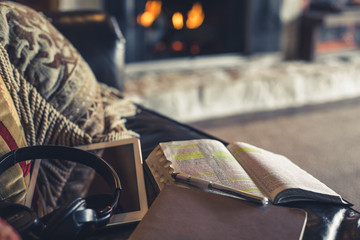 Close-up photo open Bible, coffee, notepad and tablet on couch in front of fire place