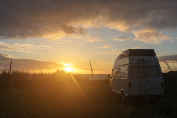 Oldtimer campervan parking at the ocean while the sun sets on the horizon casting a warm light on...