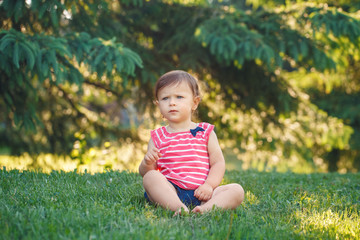 Portrait of cute adorable pensive serious little  Caucasian baby girl toddler in park outside. Happy lifestyle childhood concept. Child kid sitting on grass during summer sunset