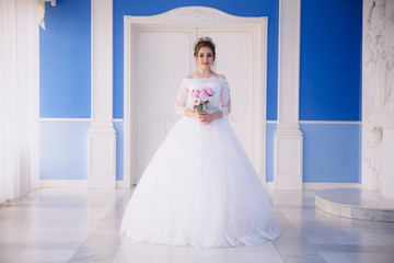 Fototapeta premium A beautiful girl in a lush white dress on her wedding day is standing with a bouquet of peonies in a blue hall with a high ceiling