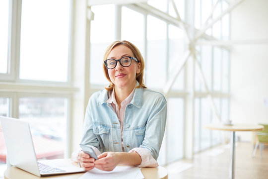 Adult charming woman in casual outfit and eyeglasses sitting at table with laptop holding phone and smiling at camera. 