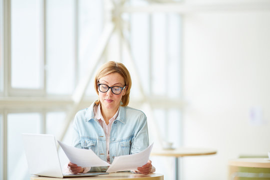 Stylish woman in glasses looking through papers and working on laptop at table in light office