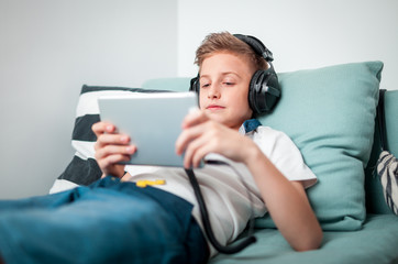 Teenage boy using tablet with headphones lying on bed in his room