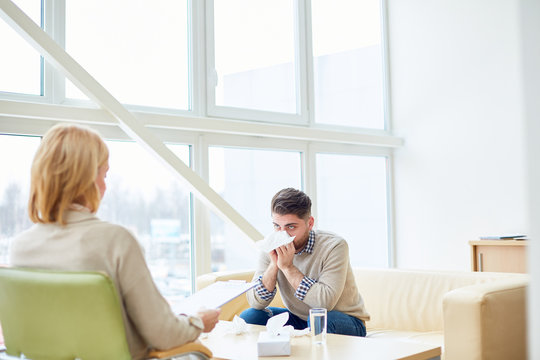 Depressed man sitting on sofa in light room and having talk with professional psychotherapist