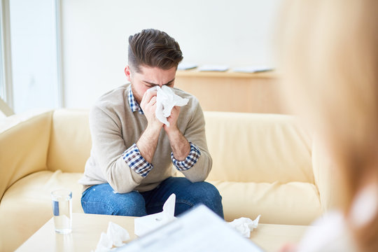 Young man blowing nose while crying on visit to psychotherapist having mental health problems