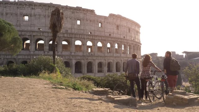 Three happy young friends tourists with bikes and backpacks at Colosseum in Rome standing on hill at sunset with trees slow motion steadycam wide shot