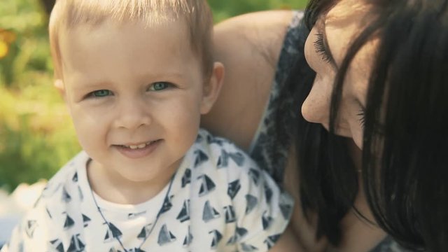 Young mother with her adorable two year old boy playing outdoors with love in slow motion. Mother kisses little son looking at camera. Concept of happy family love