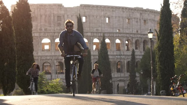 Three young friends tourists riding bikes in colle oppio park in front of colosseum on road with trees at sunset in Rome slow motion