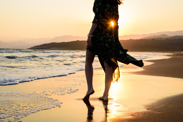 Bare feet woman in green dress on the beach before the orange sunset, silhouette with rays