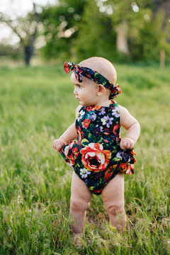 Cute one-year old girl in floral romper and headband outside in tall grass.