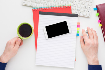 white desktop, male hands, guy holding Cup of coffee, smart phone lies on the table, guy working at the computer, red notebook, white background with copy space, for advertisement, top view