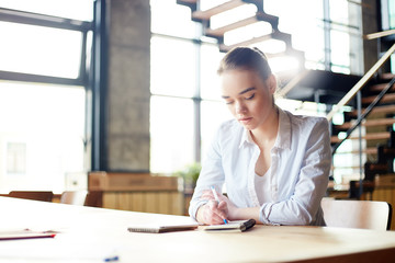 Busy young woman in shirt writing in notepad and sitting at table in daylight concentrated on...