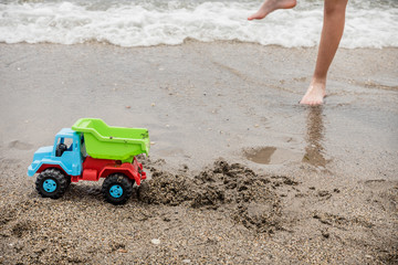 Fototapeta na wymiar green, blue and red toy truck on the seashore in front of a child's feet