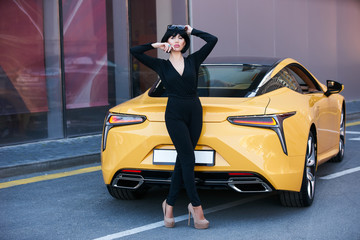 Young woman with dark hair in black bodysuit and sunglasses posing near supercar. Portrait of a...