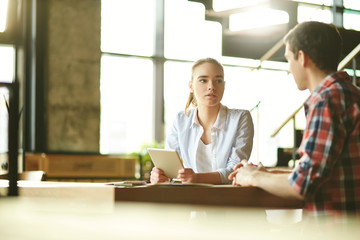 Young businesswoman with tablet in her hands and her male colleague sitting at desk and discussing