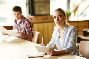 Serious confident millennial girl holding tablet, sitting at desk and looking at camera; her fellow student in background