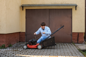Young man in jeans and shirt checks the oil level in the lawn mower opposite the garage in the summer.