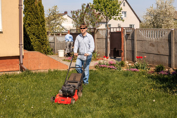 Young man in sunglasses, jeans and shirt mows the grass on the lawn in the village in the summer.