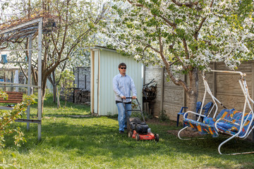 Young man in sunglasses, jeans and shirt mows the grass on the lawn in the village in the summer.